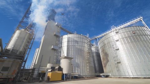 Panoramic view of agricultural plant. Large round grain elevators for storing grains. Thick fumes releasing into the air during grain processing on a factory.