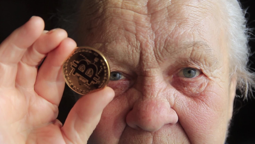 An elderly woman, a grandmother, is holding a bitcoin coin in her hands and leans it to her eye