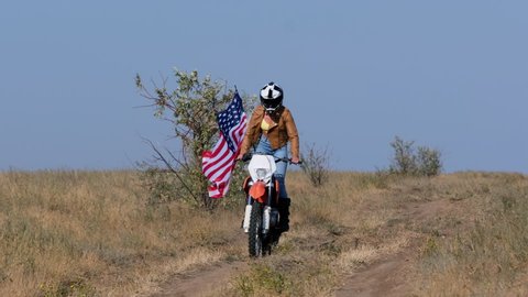 A girl rides a sports motorcycle on the ground with a waving USA flag. Off-road motorcycle competition.