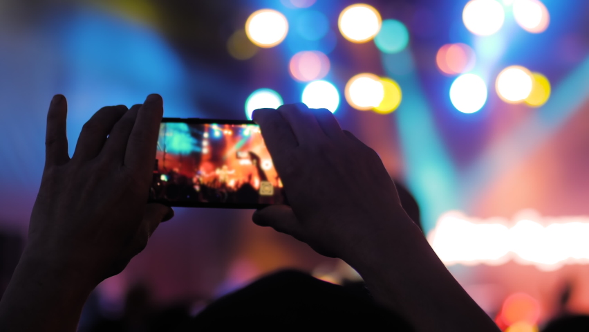 Close up: man hands silhouette taking photo or recording video of live music concert with smartphone at nightclub. Bright colorful stage lighting. Photography, technology concept