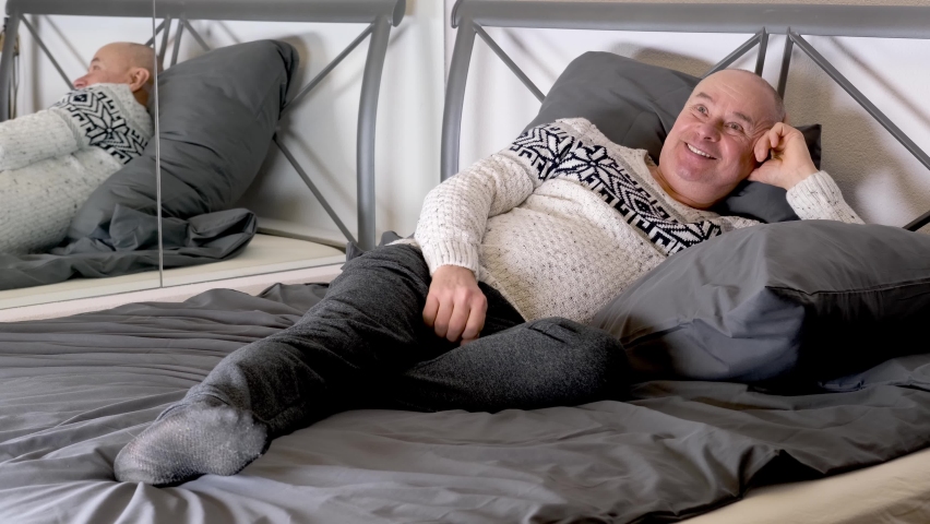 bald man in a Scandinavian pullover is lying in bed at home, resting after work, watching TV, quarantine concept, smiling happily