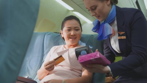 Onboard product purchasing, Asian woman sitting in airplane buying gift from air hostess and paying by credit card tapping on machine. Contactless payment and technology inflight service concept.
