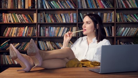 Sexy businesswoman in white blouse and yellow skirt twirls pencil drinks coffee and types on laptop putting legs on table in office against bookshelves