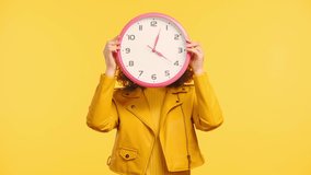 smiling, tricky woman obscuring face with round clock isolated on yellow