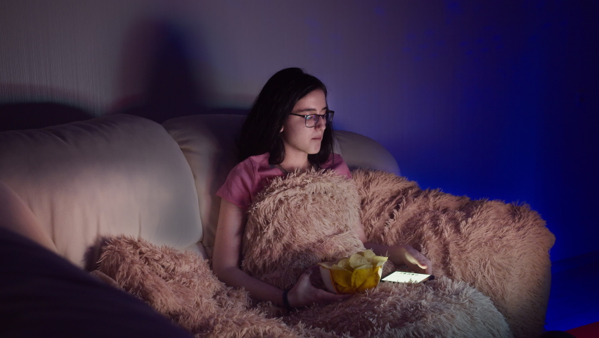 girl sitting on the sofa, watching tv uses phone and eating chips in the evening, girl relax at home