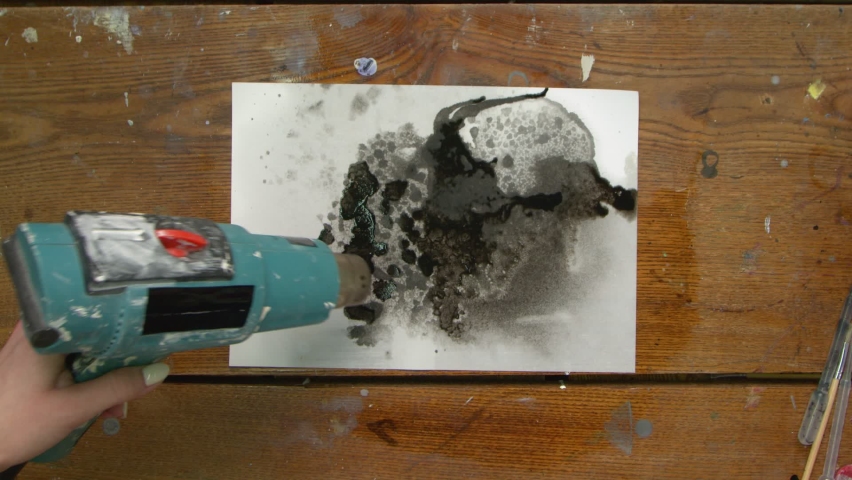 Top view of artist paints an abstract fluid picture, she uses dryer to move a blak paint on canvas and dry