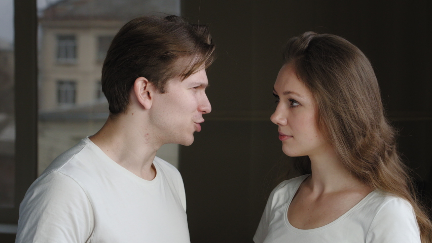 Two people couple of caucasian woman and man talking, millennial guy tells secret to wife girl, lady looking at camera surprised puts finger to male mouth forbidding to speak makes gesture of silence