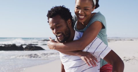African american man smiling and carrying african american woman piggyback on the beach. healthy outdoor leisure time by the sea.