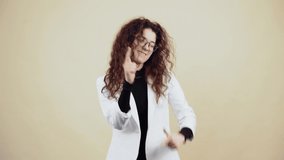 The energetic young woman with curly hair dances waving her hands, she likes music, dance moves. Young hipster in gray jacket and white shirt, with glasses posing isolated on beige background in the
