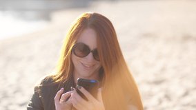 Closeup view 4k stok video footage of beautiful satisfied happy young adult woman isolated on sunny sandy beach, pleased with online communication, types text message on smartphone. Technology concept