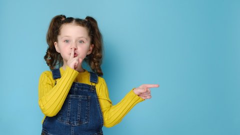 Emotional cute little girl holding finger on lips symbol of hush gesture of asking to be quiet, isolated over blue studio background with copy free space for advertisement. Silence or secret concept