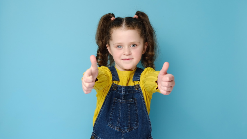 Happy beautiful smiling little preschool girl child looking at camera showing thumbs up, giving recommendation, standing isolated on blue studio background with copy space. Best life quality concept. | Shutterstock HD Video #1069792120