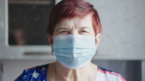 Elderly woman with short hair wearing blue medical mask standing at home and looking at camera, focus on eyes. Closeup aged person staying at home during lockdown. Concept of pandemic