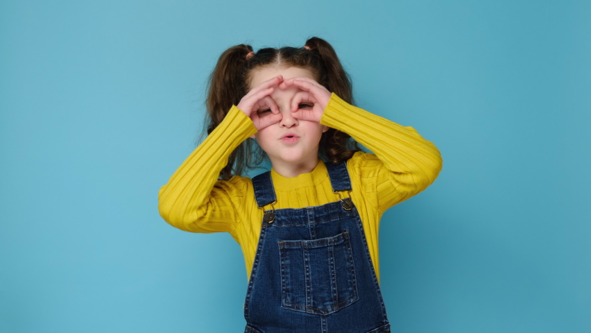 Funny playful little preschool girl making fingers like glasses eyewear shape looking through binoculars smiling looking at camera, isolated over blue studio background. Fools day and comic concept | Shutterstock HD Video #1069794022