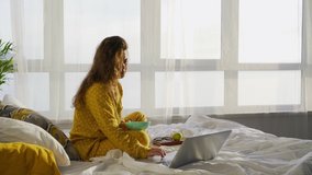 Happy young lady in yellow pajamas eats healthy breakfast watching video on laptop on comfortable bed in hotel room in lazy morning close view