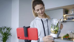 Smiling young lady food blogger in grey hoodie shows blender bowl full of cut fruits and vegetables shooting video in spacious light kitchen