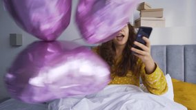 Cheerful young woman in grey mask on forehead and yellow pajamas dances with cluster of balloons and shoots video celebrating birthday in bed in morning
