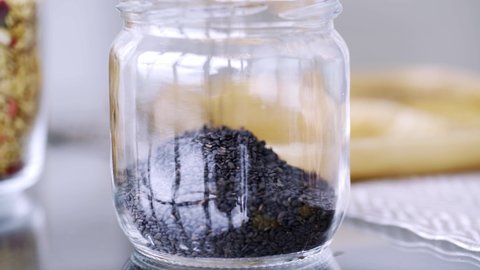 Filling transparent glass jar with small black raw sesame seeds to cook tasty organic food on shiny clean table in light kitchen extreme close view