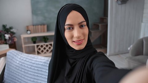 Closeup portrait pretty Asian female in black hijab posing taking selfie video call POV shot. Happy Arabic woman with natural beauty in ethnic clothes photographing at home. Shot with RED camera in 4K