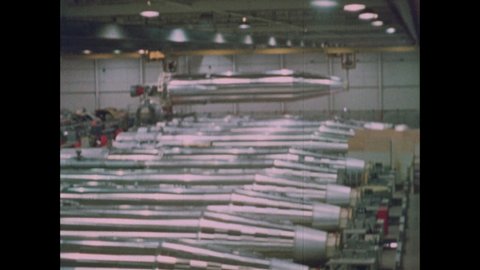1960s: ICBM Missile body is lifted and moved above a long row of other missiles and lowered into place at end of line. Crane machinery moves back in place.