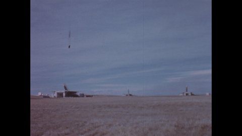 1960s: Military complex on barren ground. ICBM missile starts to raise vertically behind building. Plane flies overhead.