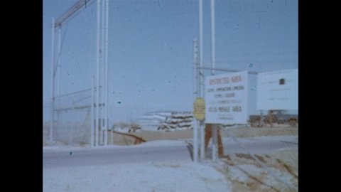 1960s: Military man stands at gate of Atlas ICBM site and waves truck through. Driver of the truck shows ID to guard. Military complex on barren ground.