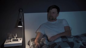 Young caucasian man suffering from depression and insomnia getting upset trying to entertain himself while watching tv via streaming service, switching channels with remote control in bed at night
