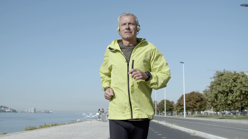Active male pensioner with hearing aid device jogging outside. Elderly man in sporty clothes keeping fit, running down street. Side view. Elderly age and lifestyle concept