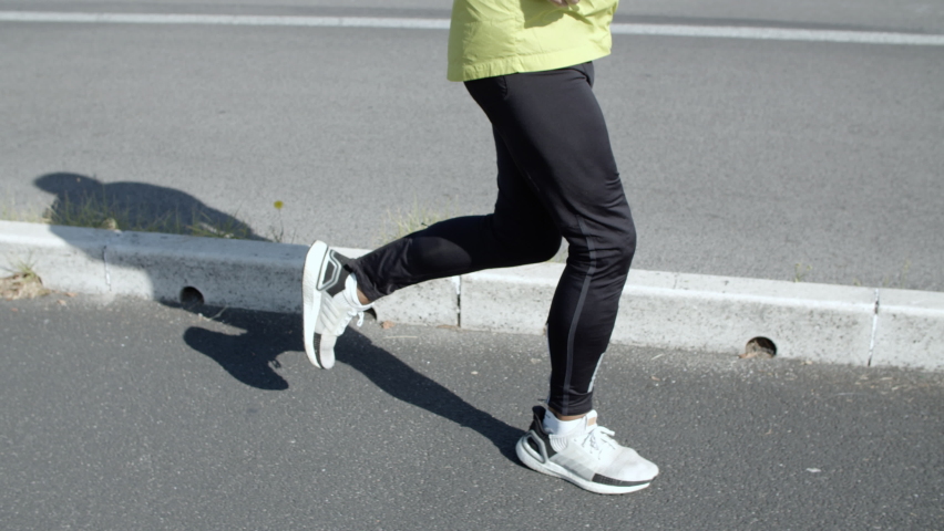 Active old jogger with headphones running outside. Elderly man in sporty clothes keeping fit, jogging down street. Dolly shot. Elderly age and lifestyle concept