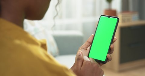 Close up of unrecognizable African American woman using smartphone with green screen and scrolling while sitting in room. Female holding black mobile phone with chroma key in hand, tech concept