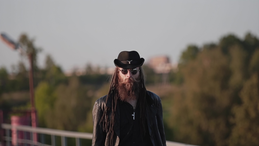 A man in a black raincoat, hat, and sunglasses walks and looks around. Nature is all around and there is no one
