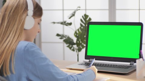 child schoolgirl at her desk does homework at home in front of green screen laptop monitor,distance education,little girl in lockdown studying from remote looking at chroma key computer display
