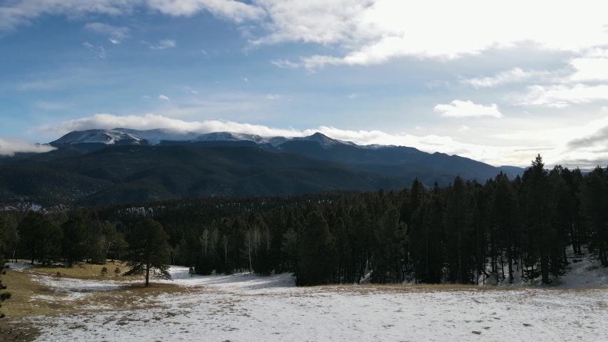 Aerial view moving forward shot, Scenic view Pine Trees Forest on the snow covered hills of Pikes Peak, Colorado Springs, Mountain range and a cloudy blue sky in the background.