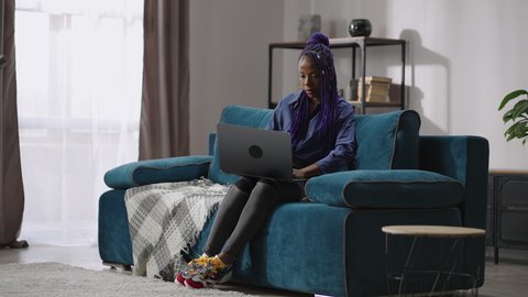 single afro-american woman is using laptop, sitting alone at home at weekend, sitting on couch in room and surfing internet, full-lenght portrait of black lady