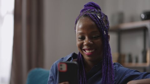 cheerful black woman is chatting by video call in smartphone and smiling broadly, laughing looking at display, resting at home, portrait of nigerian or creole lady in apartment