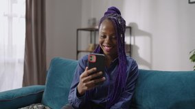 happy afro-american young woman is viewing on screen of her smartphone and smiling, communicating by video call or watching funny video in social nets, portrait shot