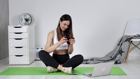 Fit woman sits on a yoga mat after an online workout. Female holding looking at her phone experiencing euphoria and joy from the results