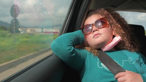 Little girl resting inside car during a trip. Child on booster seat, fasten belts wearing sunglasses and face mas.