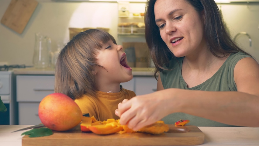 Happy Kid and mother with mango fruit in hands. Funny family crazy eating mangoes healthy dietary nutritious at home in the kitchen. Healthy lifestyle, raw food Royalty-Free Stock Footage #1069823908