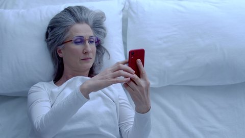 Mature female yawning, putting smartphone on pillow, taking eyeglasses off, rest
