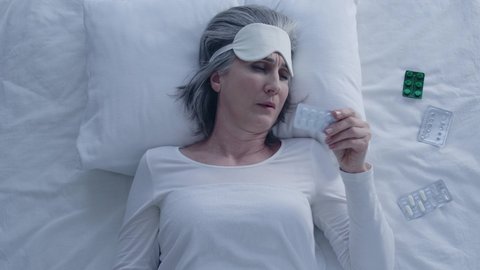 Senior greyhaired woman taking sleeping pills in bed putting on eye mask, health
