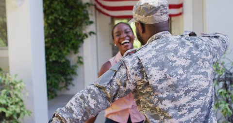 African american male soldier hugging smiling wife in front of house with american flag. soldier returning home to family.