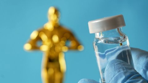 Hollywood Golden Oscar Academy award statue and hand in gloves with vaccine aganist coronavirus on blue background. Success and victory concept. Oscar ceremony in coronavirus time concept