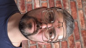 Vertical video. The bearded man put several glasses on his face. Short haircut. Blue T-shirt. Poor eyesight. Choice of glasses. Optics. Against the background of a brick wall. Large portrait.