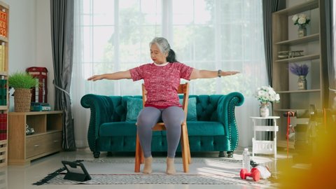 Handheld slow motion shot, Senior woman sitting on chair to exercise shoulders and arms, Training exercise online with tablet In Living Room During Quarantine