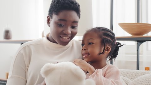 Afro american mother with little daughter girl sitting on sofa at home talking holding teddy bear. Single parent ethnic black mom mixed race elder sister woman and child female kid chatting indoors 스톡 비디오