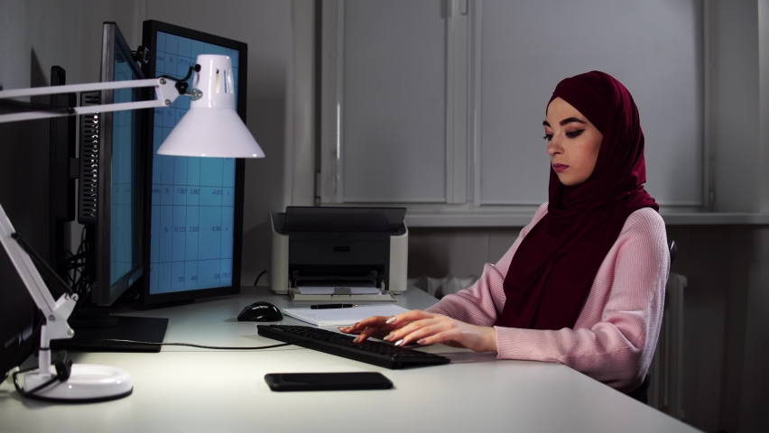 A woman in hijab sits at office in front of computer and types on keyboard. She prints document with graphs and shows it by videocall. Young muslim lady does her job distantly using virtual technology
