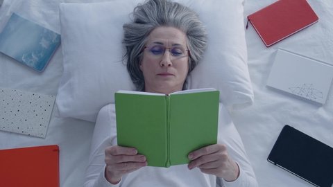 Greyhaired woman in eyeglasses reading in bed, lying on pillow, feeling lonely