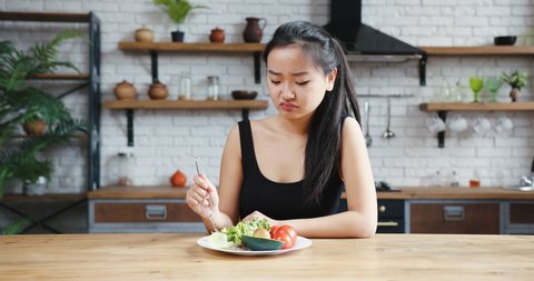 Asian woman sitting at table, looking sad and bored with diet not wanting to eat salad. Modern kitchen interior
