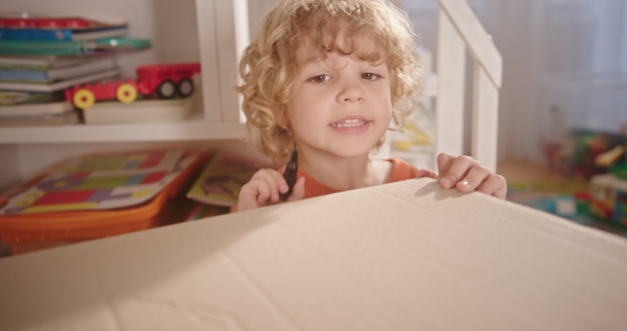 Cute boy drawing on carton box. Tracking shot of adorable boy with curly hair talking and drawing on cardboard box at home
